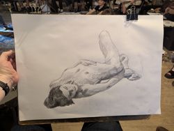 A pencil sketch of a woman lying down, her head towards you. In the background of the photo is the drawing studio with other artists